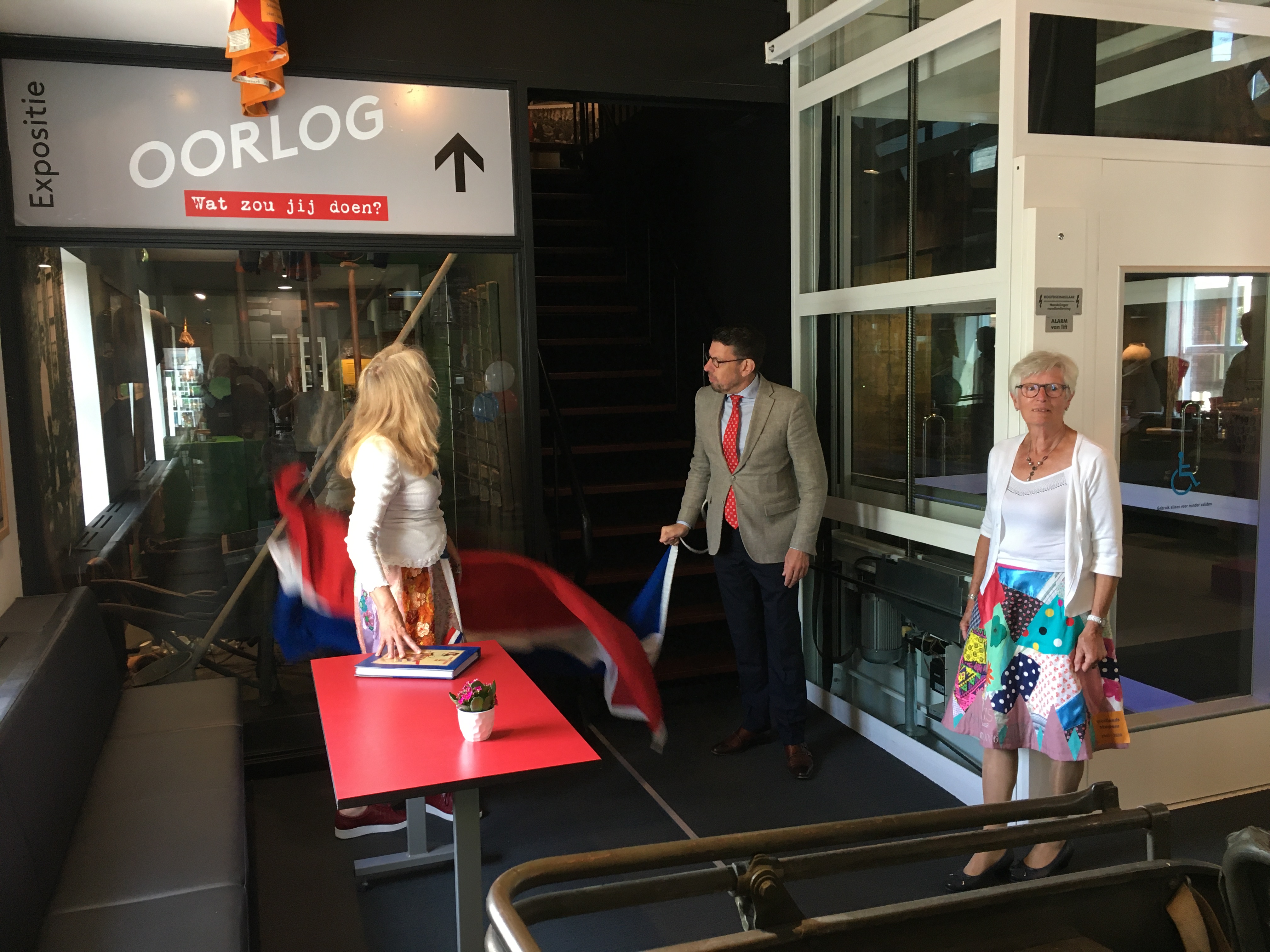 Burgemeester Arends opent - begeleid door vrijwilligers in bevrijdingsrok - de expositie door de vlag weg te trekken