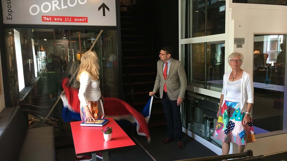 Burgemeester Arends opent - begeleid door vrijwilligers in bevrijdingsrok - de expositie door de vlag weg te trekken